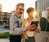 A man and a woman look together at a tablet outdoors at sunset.