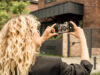 Una donna dai riccioli biondi sta fotografando con il suo smartphone un moderno edificio residenziale in mattoni.