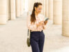 Una donna sorridente con gli occhiali sta usando il suo smartphone davanti a un edificio moderno con colonne.